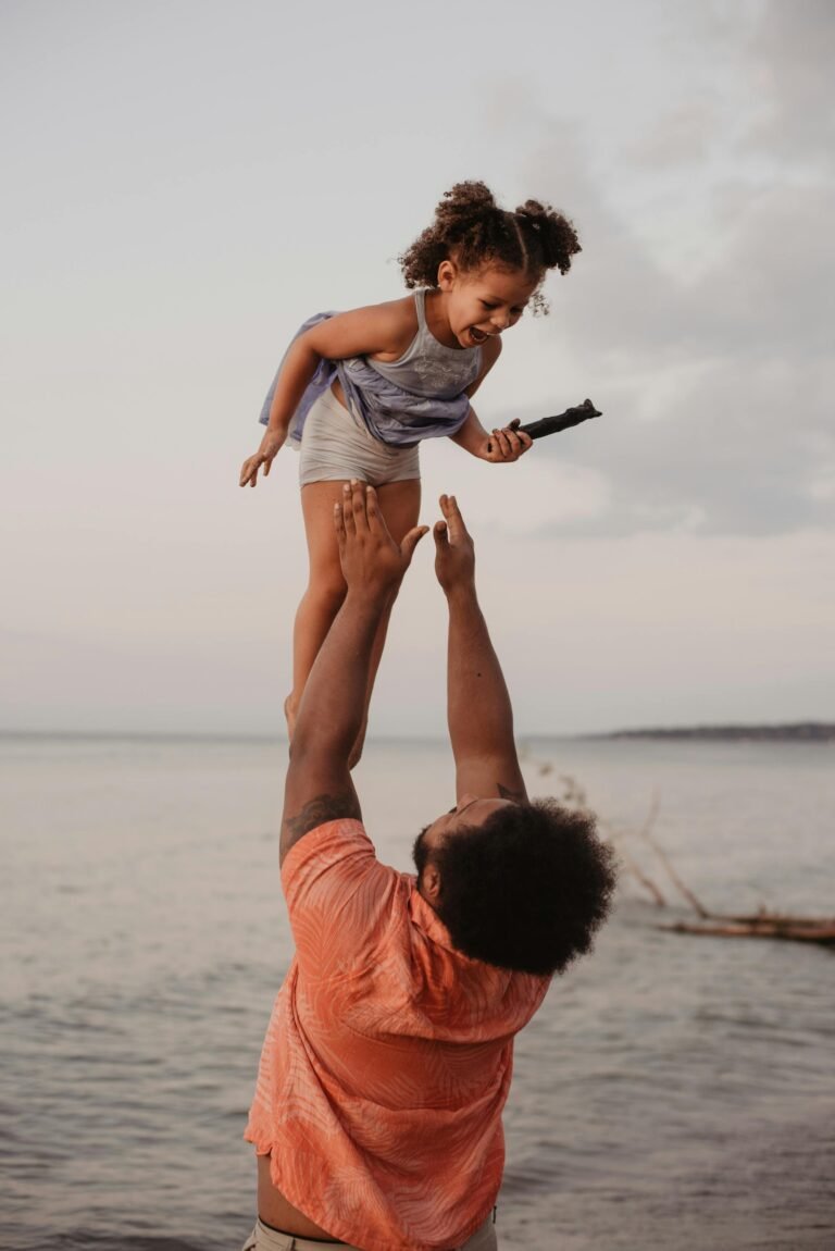A joyful father lifts his daughter in the air by the beach during sunset, capturing a moment of love and happiness.